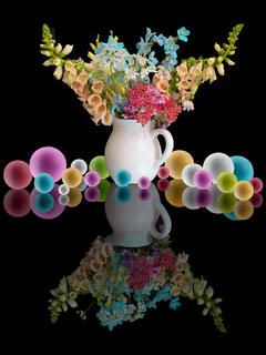 A fine art portrait-orientation still life photograph on a black background featuring a white ceramic pitcher filled with a lush wildflower bouquet