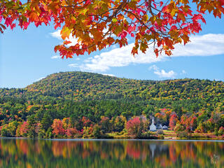 Vibrant red and orange autumn maple leaves frame a scenic New England lake reflection with a white church and rolling foliage-covered hills under a blue sky.