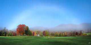 A wide panoramic New England autumn scene featuring a rare white fogbow arching across a clear blue sky, above a lush green field with a vivid red maple tree