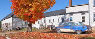 Wide panoramic view of a rusty vintage 1950s blue and white two-tone car parked alongside a large white New England farmhouse complex with antique farm equipmen
