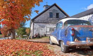Close-up rear view of a rusty vintage 1950s blue and white two-tone car parked in front of a tall weathered gray New England barn with cupola and weather vane, 