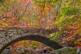 A curved mossy granite stone arch bridge over a rocky woodland stream, surrounded by dense red, orange, yellow, and green autumn foliage trees and vines in NH