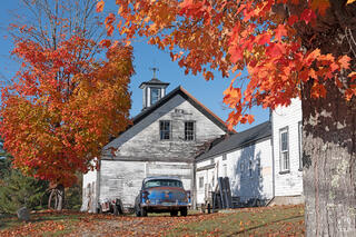 A vintage 1950s blue classic car parked beside a large weathered white New England barn with a cupola and weather vane, framed on both sides by brilliant orange