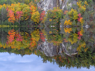 Colorful red, orange, yellow, and green autumn trees growing alongside a dramatic granite rock cliff, perfectly reflected in a calm still lake with soft cloudy