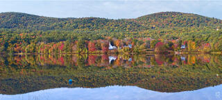 Wide view of twin rolling hills covered in red, orange, and green fall foliage reflected in a calm New England lake, with a white church steeple, nearby homes, 