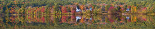 Ultra-wide panoramic view of peak New England fall foliage in red, orange, yellow, and green reflected in a glassy lake, with a white church steeple and houses 